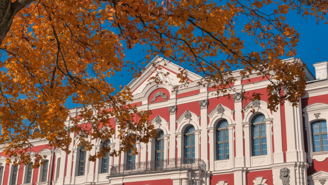 Latvijas Biozinātņu un tehnoloģiju universitātes ēka - klasiskā stila pils sarkanā krāsā ar baltiem logu rāmjiem un kolonnām. Priekšplānā redzam nelieks klavas zars ar rudenīgām, dzeltenām lapām