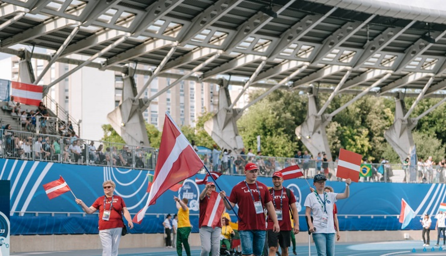 Latvijas paralimpiskās komandas pārstāvji pārvietojas pa stadionu ar Latvijas karogiem rokās
