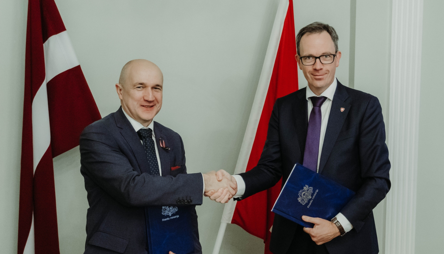Uz Latvijas un Šveices karoga fona Šveices Konfederācijas vēstnieks Martēns Mišlē un Finanšu ministrijas valsts sekretāres vietnieks ES fondu jautājumos Armands Eberhards spiež viens otram roku