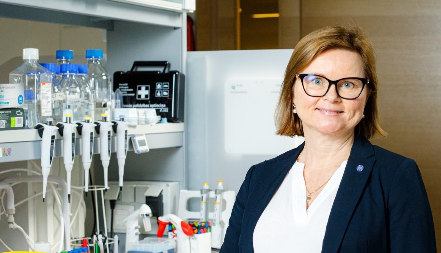Una Riekstiņa priecīga stāv pie laboratorijas galda, uz galda dažādi laboratorijas trauki – konteineri