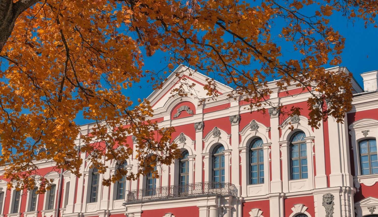 Latvijas Biozinātņu un tehnoloģiju universitātes ēka - klasiskā stila pils sarkanā krāsā ar baltiem logu rāmjiem un kolonnām. Priekšplānā redzam nelieks klavas zars ar rudenīgām, dzeltenām lapām