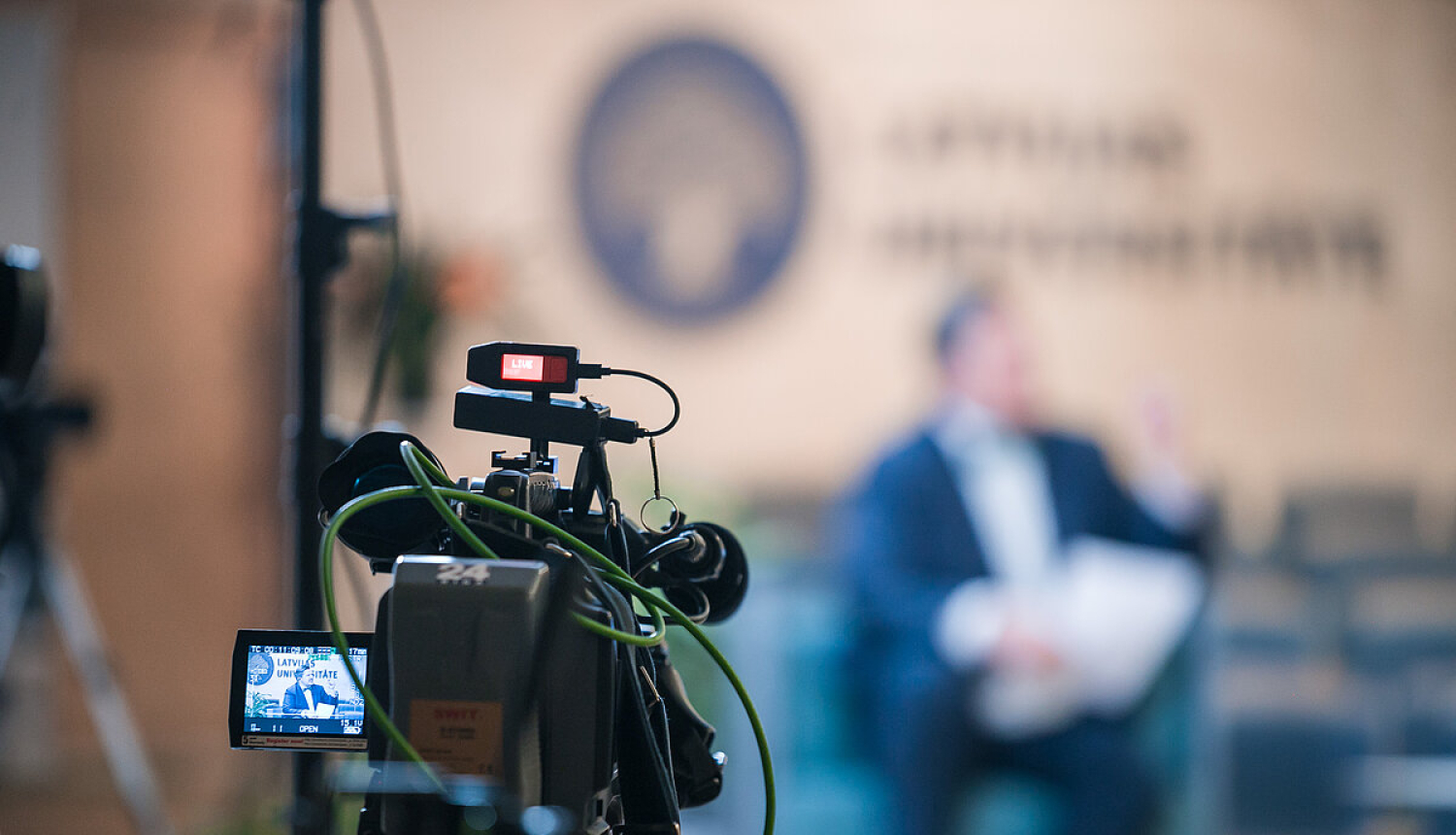 Priekšplānā video kamera, kas filmē runātāju, kurš sēž telpā. Uz sienas redzams Latvijas Universitātes logo. 