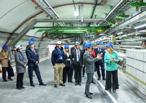 CERN pārstāvji stāsta un rāda telpas premjerministrei Evikai Siliņai un citiem Latvijas delegācijas pārstāvjiem. Visi atrodas industriālās telpās ar zilām ķeverēm galvās.