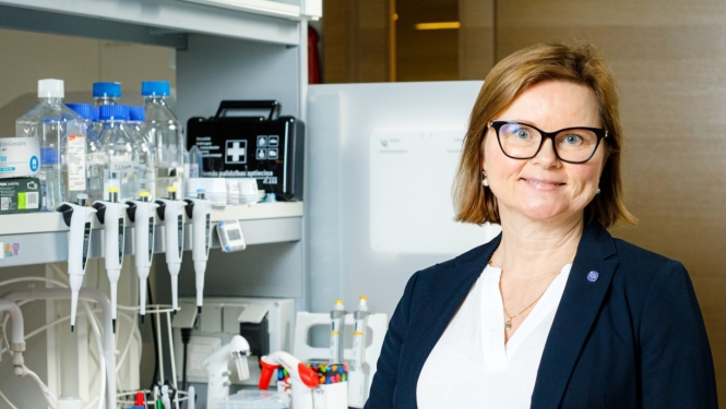 Una Riekstiņa priecīga stāv pie laboratorijas galda, uz galda dažādi laboratorijas trauki – konteineri