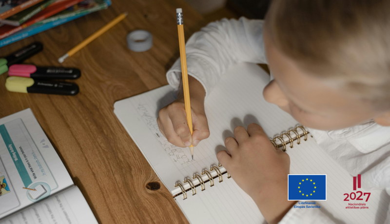 Bērns raksta ar zīmuli kladē, kadrs uzņemts no augšas, redzams galds. Apakšējā labajā stūrī redzams Eiropas savienības un Nacionālā attīstības plāna logotipi