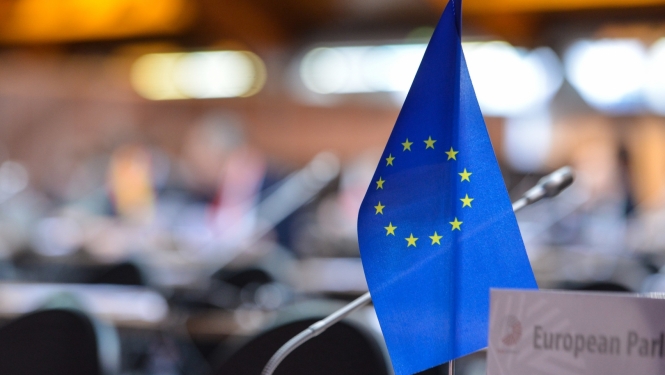 Priekšplānā Eiropas Savienības karodziņš, aizmugurē konferences telpas sēdvietas