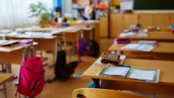 Klase, uz galdiem stāv mācību grāmatas un burtnīcas. Pie galdiem karājas skolas mugursomas.