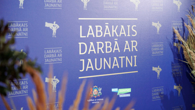 "lABĀKAIS DARBĀ AR JAUNATNI" stends, apkārt stāv augi