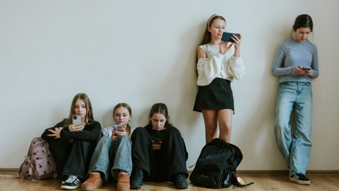 Skolēni atspiedušies pret sienu un apsēdušies uz zemes, pēta telefonus
