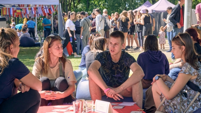 Jaunieši sēž un sarunājas, fonā daudz cilvēku un telts ar dažādu valstu karodziņiem