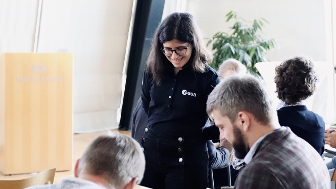 Smaidīga meitene stāv blakus galdam, kur sēž divi smaidīgi vīrieši. Notiek mācību process.