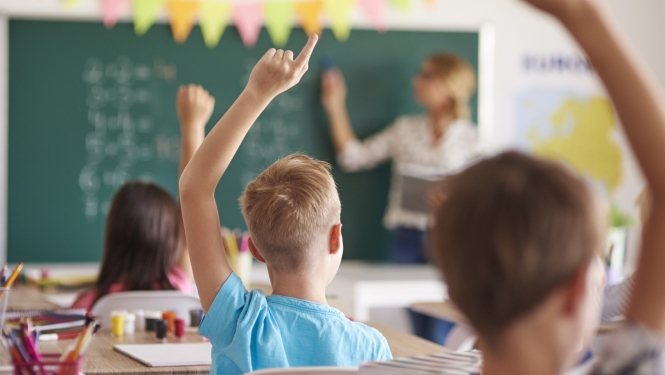 Bērni sēž klasē un ceļ roku, priekšā pie tāfeles stāv skolotāja