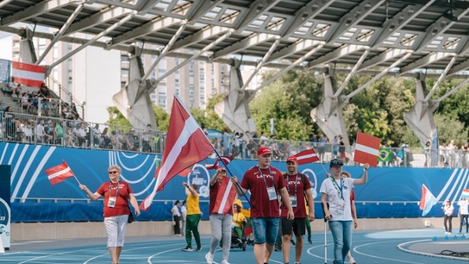 Latvijas paralimpiskās komandas pārstāvji pārvietojas pa stadionu ar Latvijas karogiem rokās