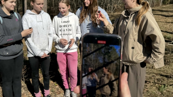 Piecas jaunietes stāv un sarunājas ārā, aizmugurē koki, priekšā kāds filmē šo sarunu tiešraidē