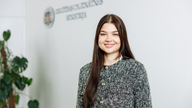 Silvija Amatniece stāv uz gaišas sienas fona. Uz sienas ir uzraksts "Izglītības un zinātnes ministrija"
