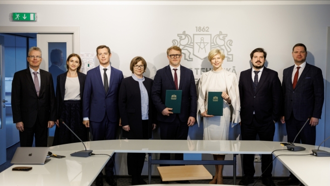 RTU pārstāvju un ministrijas pārstāvju kopbilde pie sienas, uz kuras redzams liels RTU logo