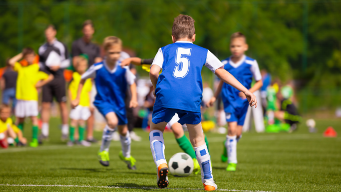 Puikas zilās formās spēlē futbolu. Fonā stāv citi puikas dzeltenās formās un treneri.