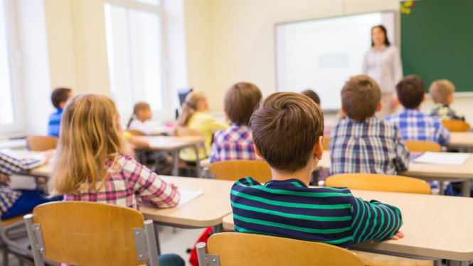 Bērni sēž klasē, skats no mugurpuses, priekšplānā puika strīpainā džemperī un meitene rūtainā kreklā. Tālumā klases priekšā ir skolotāja. 