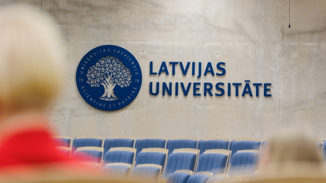 Latvijas Universitātes auditorija ar ziliem krēsliem, uz sienas liels zils LU logotips - ozols zilā aplī un uzraksts Latvijas Universitāte