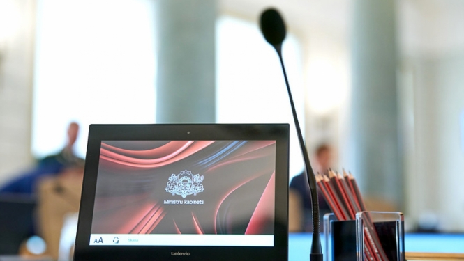 Dators un mikrofons stāv uz galda Ministru kabineta zālē, datora ekrānā redzams Ministru kabineta logotips