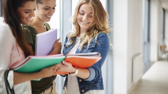 Trīs studentes stāv un smaida, rokās tur mācību klades