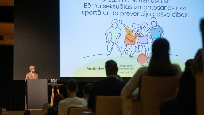 Izglītības un zinātnes ministre Anda Čakša konferencē uz skatuves, aiz viņas liels ekrāns ar bērnu ilustratīvu zīmējumu