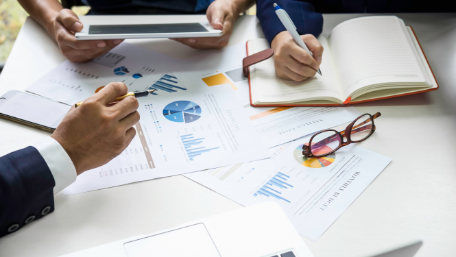 Uz galda noliktas papīra lapas ar grafikiem un diagrammām, redzamas trīs cilvēku rokas - viens cilvēks tur planšeti, otrs raksta kladē, trešais rāda ar pilspalvu uz diagrammu