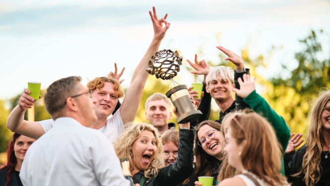 Grupa jaunu cilvēku svin pasākuma norisi brīvā dabā, smaidot un priecājoties. Atmosfēra ir dzīvespriecīga un draudzīga, fonā redzami koki un gaišas debesis.