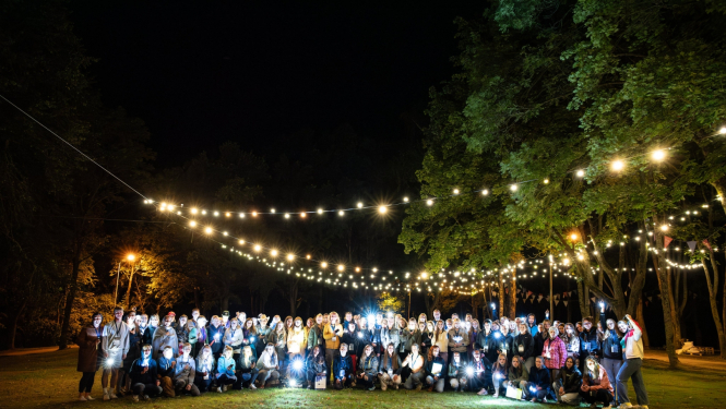 Projektu nakts 2023. dalībnieku kopbilde, vakarā, kad debesis ir tumšas, virs dalībniekiem iekārtas gaimas lampiņu virtenes