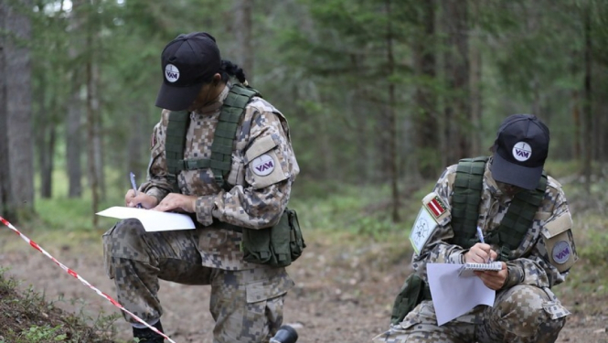 Cilvēki karavīru formas tērpos, veic pierakstus uz lapas dabā