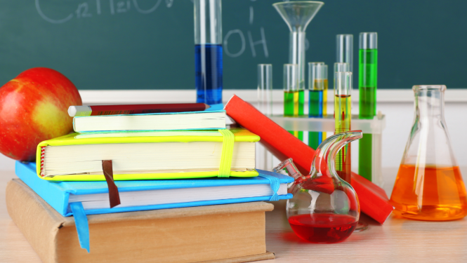 Galds klasē ar grāmatām, ābolu, kolbām, kurās ir dažādu krāsu šķīdumi. Fonā tāfele, uz kuras uzrakstītas ķīmijas formulas.