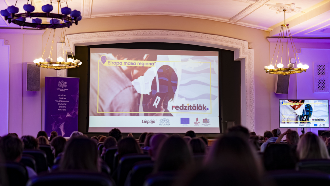 Pasākuma "Eiropa manā skolā" titulbilde uz lielā ekrāna RTU Liepāja lielajā zālē, zāle pilna ar pasākuma dalībniekiem