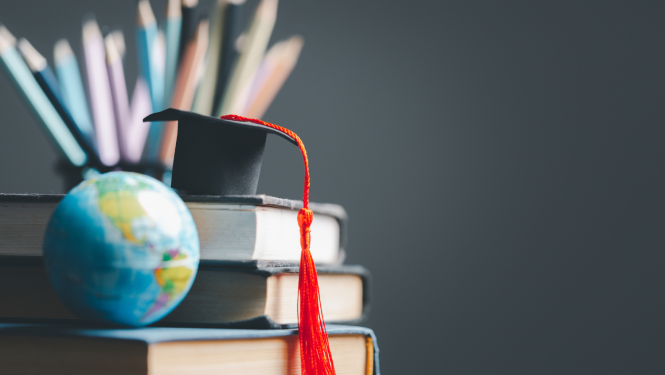 Absolventa cepure novietota uz grāmatām, blakus stāv globuss, fonā trauks pilns ar zīmuļiem.