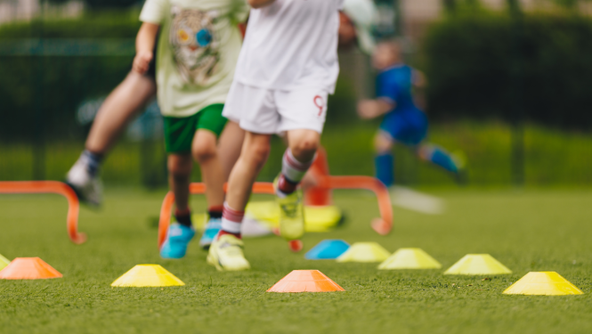 bērni spēlē futbolu, uz laukuma novietoti šķēršļi.