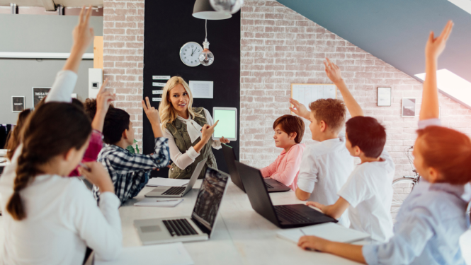 Skolēni stāv pie datoriem pacēluši rokas. Sieviete rāda uz skolēniem.