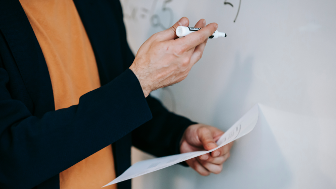 Pedagogs tur rokās darba lapu un rakstāmo, rāda uz tāfeli.