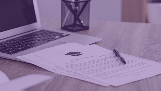 Uz galda stāv dators, dokumenti un pildspalva, uz dokumenta attēlota absolventa cepurītes ikona