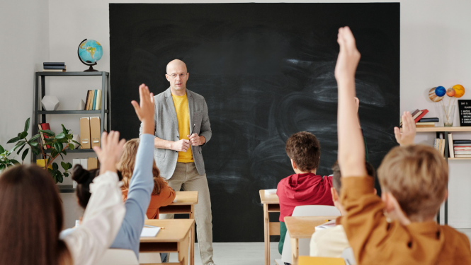 Skolotājs stāv pie tāfeles un skatās uz bērniem. Bērniem paceltas rokas.