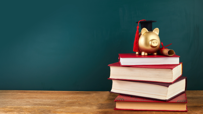 uz galda stāv mācību grāmatas, uz tām stāv krājkasīte ar uzvilktu absolveta cepurīti