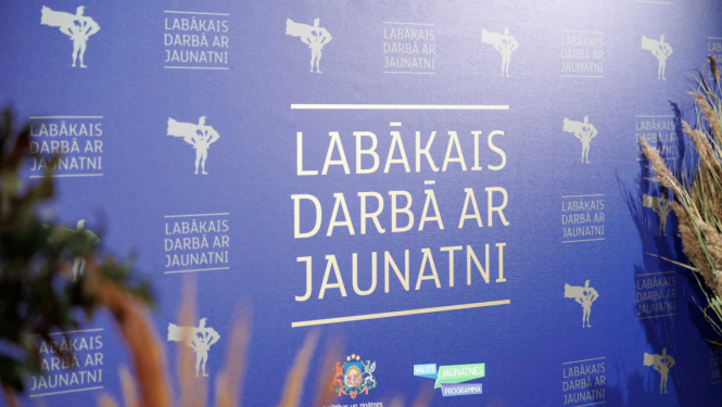 "lABĀKAIS DARBĀ AR JAUNATNI" stends, apkārt stāv augi