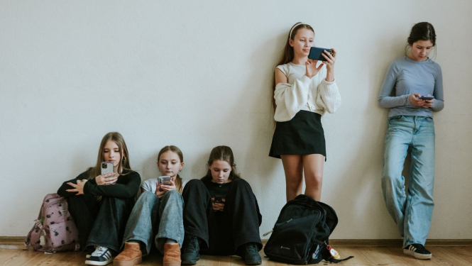 Skolēni atspiedušies pret sienu un apsēdušies uz zemes, pēta telefonus