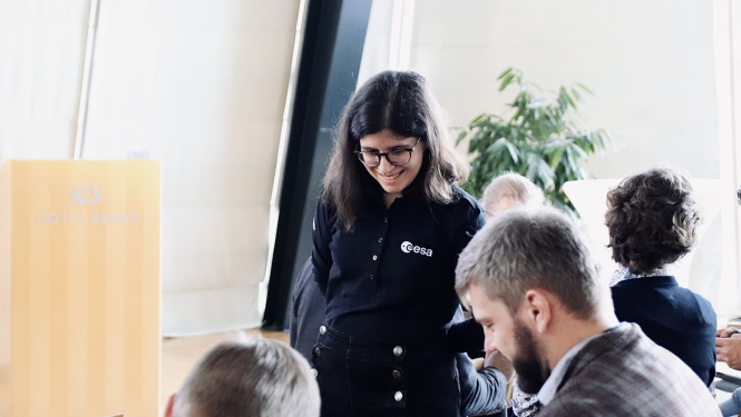 Smaidīga meitene stāv blakus galdam, kur sēž divi smaidīgi vīrieši. Notiek mācību process.