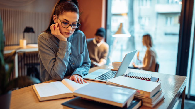 Meitene sēž pie galda ar grāmatu, uz galda noliktas grāmatas un dators