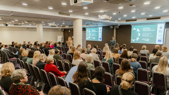 Konferece norises vietā cilvēki sēž krēslos, priekšā runātāja stāsta, fonā lieli ekrāni.