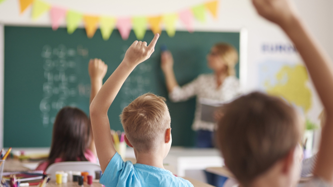 Bērni sēž klasē un ceļ roku, priekšā pie tāfeles stāv skolotāja