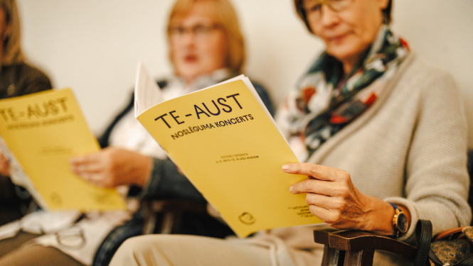 Sievietes tur rokās bukletus ar uzrakstu "TE-AUST noslēguma koncerts"