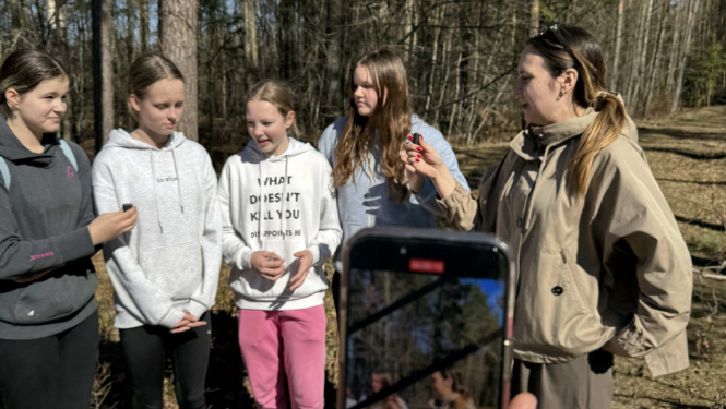 Piecas jaunietes stāv un sarunājas ārā, aizmugurē koki, priekšā kāds filmē šo sarunu tiešraidē