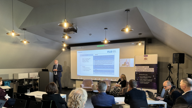 Foto no semināra, vīrietis stāsta, citi cilvēki sēž pie galdiem