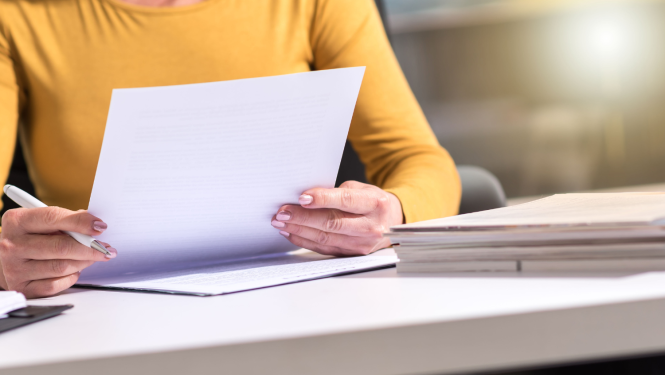 Sieviete tur vienā rokā dokumentu, otrā pildspalvu. Uz galda stāv vairāki dokumenti.