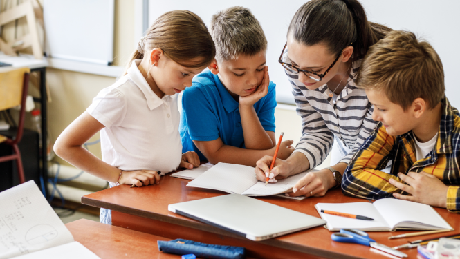 Skolotāja raksta ar zīmuli burtnīcā, apkārt stāv trīs bērni un skatās, ko skolotāja raksta